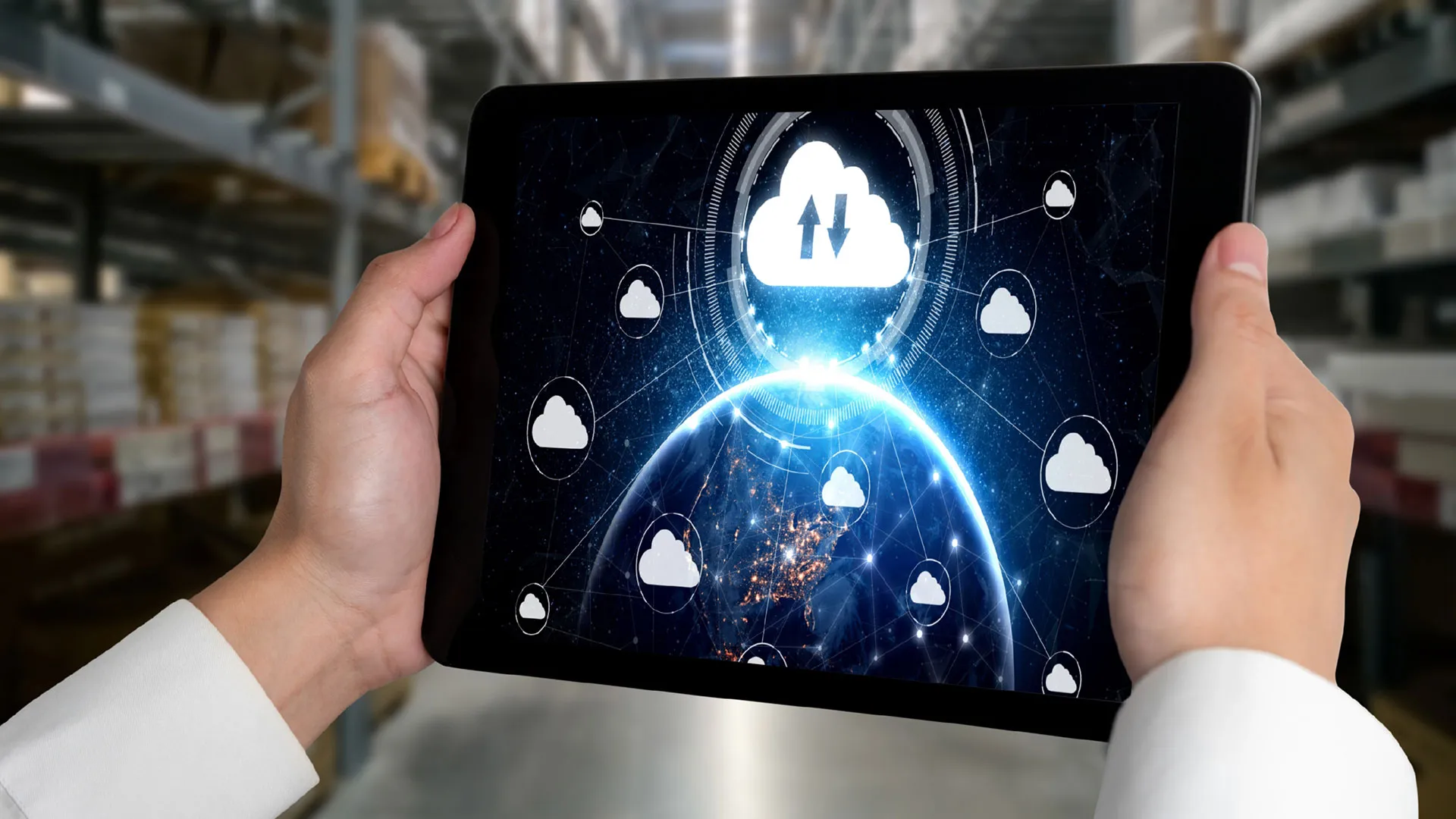 Hands holding a tablet displaying a connected cloud network over a globe, in a warehouse